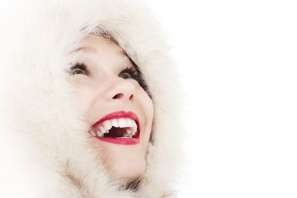 Woman on a white backdrop wearing a white fur lined coat with a hood. She's looking up in joy, and smiling. Her teeth are white which is highlighted by her red lips.