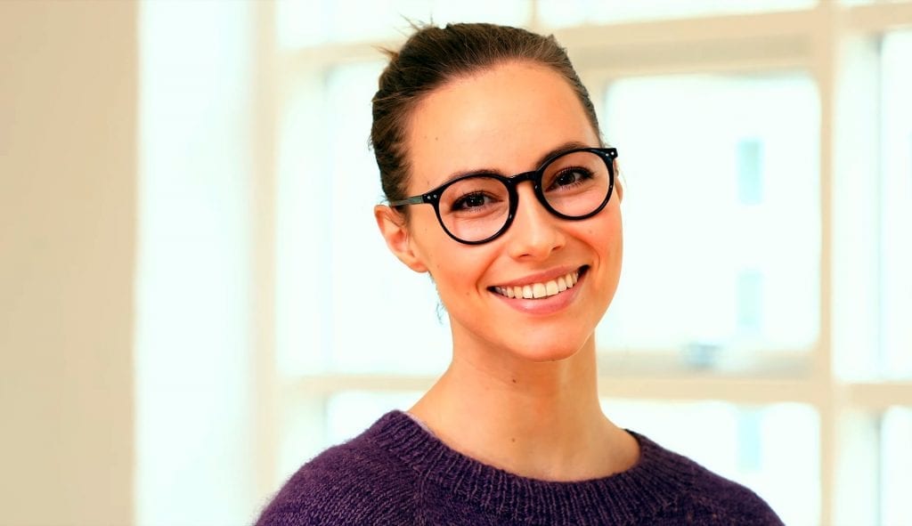 Brunette woman wearing black rim circle glasses with white teeth smiling at the camera in front of a window in a white room